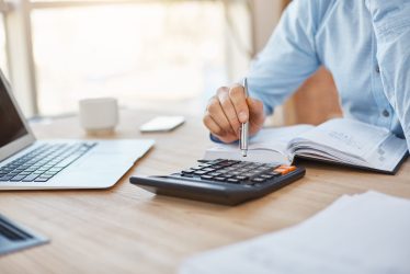 Close up detail of professional serious accountant sitting in light office, checking company finance profits on calculator, writing down results in notebook. Business concept